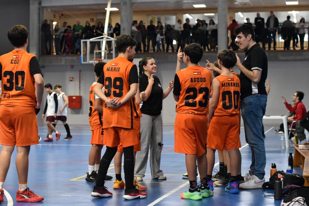 Jugadores de un equipo de cantera de Vallekas Basket atienden a su entrenadora y a su entrenador durante un tiempo muerto, en el lateral de la pista y con el público al fondo.